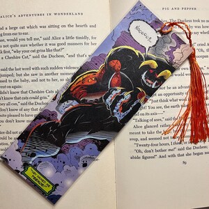 May include: Comic book-style bookmark featuring a menacing, horned creature with sharp claws and the text "THEY NAMED ME TREMOR!" and "Nooooo!!..." in speech bubbles. The bookmark has an orange tassel and is set against the backdrop of an open book.