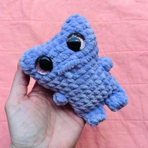 May include: A small, light blue plush toy made of soft, textured yarn. The creature features two large, round, black safety eyes with subtle pink glitter accents around the edges. It has a simple, rounded body shape with small limbs and a tiny, light blue stitched mouth detail.