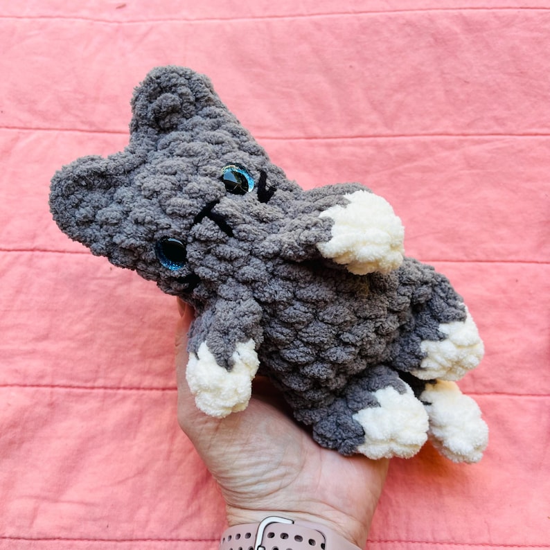 May include: A small, handmade crochet cat plush toy made from soft, textured grey yarn. The cat features white paws, a white chest patch, and two bright blue plastic safety eyes. The ears are pointed, and the overall construction is plush.
