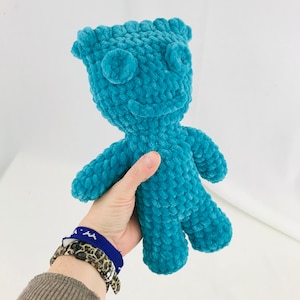 May include: A teal blue crocheted plush toy in the shape of a monster with a smiling face. The toy is being held by a hand.