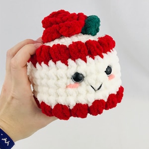 Op de afbeelding: Een gehaakt pluche speelgoed in de vorm van een taart met rood en wit glazuur en een rode roos erop. Het speelgoed heeft een glimlachend gezicht met zwarte ogen en een rode neus.