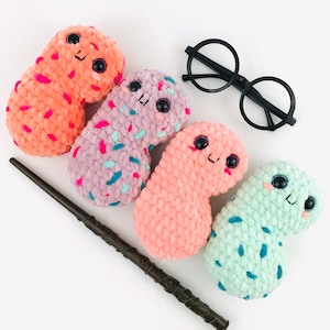 May include: Four crocheted bean-shaped plushies with different colors and patterns. The plushies have embroidered eyes and smiling faces. A black pair of glasses and a brown wooden wand are also in the image.