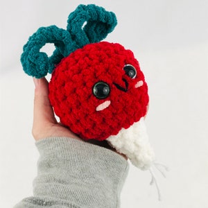 May include: A crocheted radish plush toy with a red body, white root, and green leafy top. The toy has large black eyes and a smiling face.