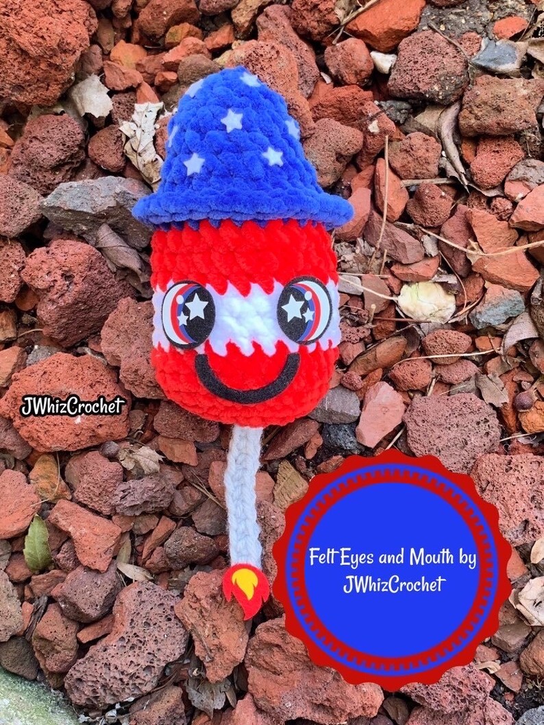 May include: A red, white, and blue crocheted firework with felt eyes and a mouth. The firework is wearing a blue and white crocheted hat with white stars. The caption reads "Felt Eyes and Mouth by JWhizCrochet".