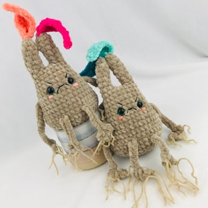 Puede incluir: Dos juguetes de peluche de crochet con forma de conejos con extremidades largas y delgadas y colas de hilo. El conejo de la izquierda tiene detalles de hilo rosa y turquesa, mientras que el conejo de la derecha tiene detalles de hilo turquesa. Ambos conejos tienen ojos y bocas bordados.