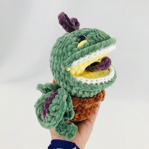 May include: Green crocheted plush toy shaped like a plant with a large open mouth and a purple tongue. The toy is in a brown crocheted pot.