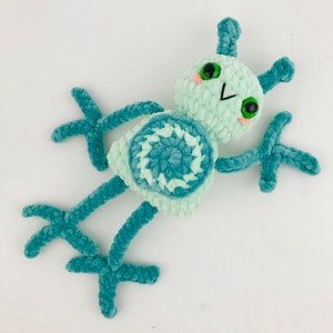May include: A teal blue and light green crocheted plush toy with a circular design on its body and large green eyes. The toy has long, thin arms and legs.