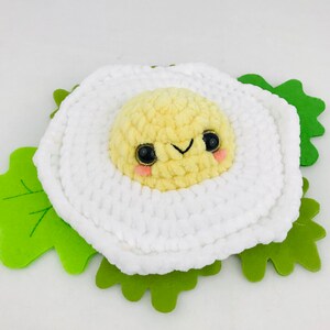 Puede incluir: Un juguete de peluche de ganchillo con forma de huevo frito con una cara sonriente. El huevo es amarillo y blanco y est&aacute; sentado sobre una hoja de fieltro verde.