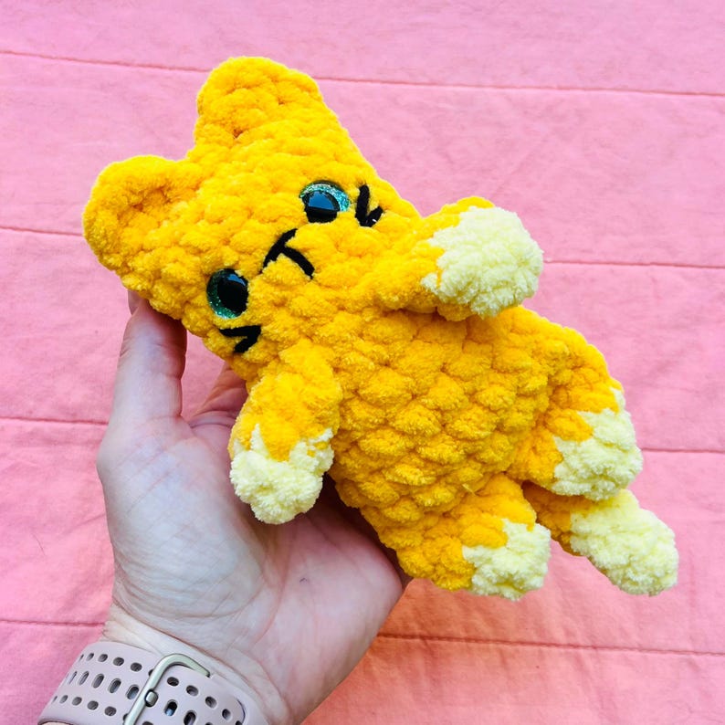 May include: A small, handmade crochet plush toy shaped like a cat, crafted from bright yellow yarn with lighter cream-coloured accents on the paws and ears. The cat features large, dark, glittery safety eyes and simple black stitched facial details.