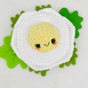 Puede incluir: Un juguete de peluche blanco de crochet con forma de huevo frito con una yema amarilla y una cara sonriente. El huevo est&aacute; rodeado de hojas de fieltro verde.
