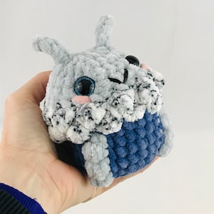 Può includere: Un piccolo giocattolo di peluche all'uncinetto grigio e blu con grandi occhi neri e un naso rosa. Il giocattolo ha la forma di una creatura con un orecchio lungo e appuntito.