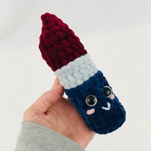 以下が含まれることがあります： 赤、白、青のカラーリングでリップスティックの形をしたぬいぐるみ。おもちゃは黒い目とピンクの頬で笑顔をしています。