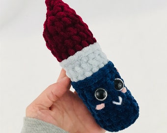 May include: A plush toy shaped like a lipstick with a red, white and blue colour scheme. The toy has a smiling face with black eyes and pink cheeks.