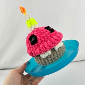 May include: A pink and grey crocheted cupcake with white and green frosting and a yellow and orange candle on top. The cupcake is sitting on a blue plastic plate.