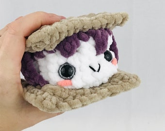 May include: A crocheted plush toy shaped like a s'more. The marshmallow is white with black eyes and pink cheeks. The chocolate is purple and the graham crackers are brown.