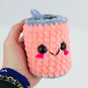 Könnte beinhalten: Ein gehäkeltes Plüschtier in Form einer Dose mit rosa Körper, grauem Deckel und einem lächelnden Gesicht mit schwarzen Augen und rosa Wangen.