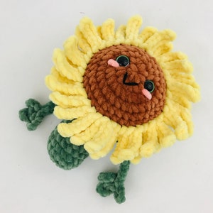 May include: A crocheted sunflower plush toy with a brown face, green stems, and yellow petals. The sunflower has a happy expression with black eyes and pink cheeks.