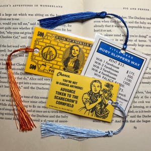 May include: Bookmarks with colorful tassels featuring designs from the Wizard of Oz and Monopoly. One bookmark is yellow with a picture of Dorothy and the Scarecrow. Another is a yellow $500 bill. A third is a Ruby Slippers Way property card.