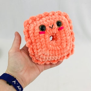 May include: A crocheted donut-shaped plush toy with a pink and orange color scheme. The toy has black eyes and pink cheeks.