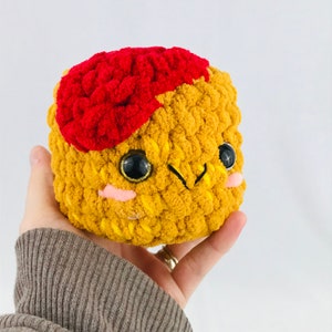 May include: A handmade, plush toy with a smiling face. The toy is primarily yellow with a red patch on top. It has large, black eyes with gold accents and small pink cheeks. The toy is held in a hand.