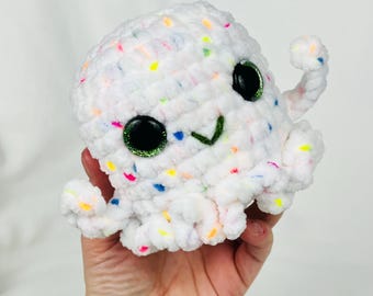 May include: A white crocheted octopus plushie with colorful speckles. The octopus has large, black, glittery eyes and a green stitched smile. Its tentacles are curled, and it appears to be held by a person.