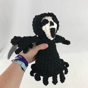 May include: A crocheted plush toy of the Ghostface killer from the Scream movie franchise. The toy is black with a white mask and a gray knife.