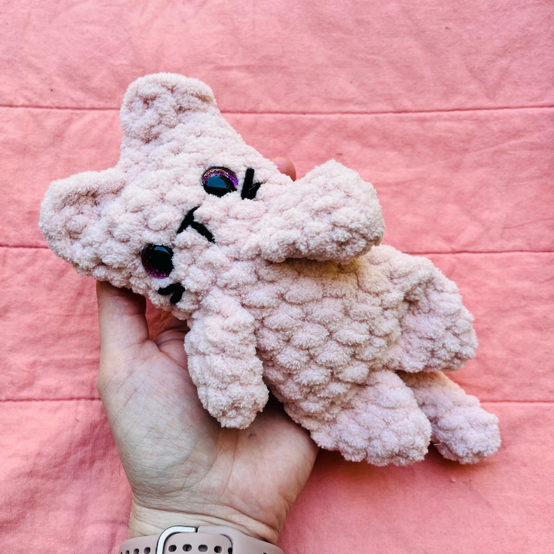 May include: A small, light pink crocheted plush toy shaped like a cat. The toy features a textured, fuzzy yarn surface, two pointed ears, and two dark, glossy safety eyes. A simple black stitched nose and mouth are visible on the centre face.