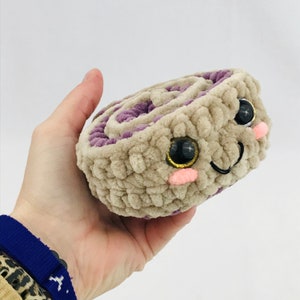 May include: A crocheted cinnamon roll plush toy with a purple swirl filling. The toy has a smiling face with large black eyes, pink cheeks, and a black stitched smile.