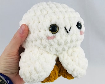 Puede incluir: Juguete de peluche blanco tejido a crochet con pies marrones y grandes ojos negros. El juguete tiene una expresión linda y de tipo caricatura.