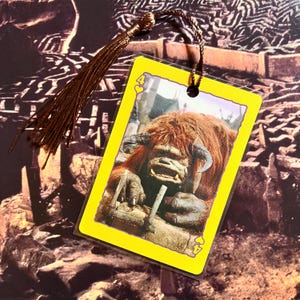 May include: A bookmark featuring a character from a fantasy film. The bookmark has a yellow border and a brown tassel. The image on the bookmark is of a horned creature with red fur. The card shows the number 4 and a heart symbol.