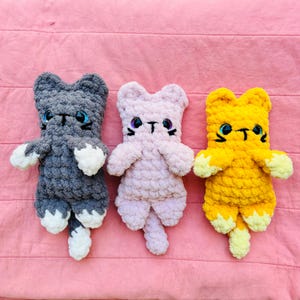 May include: Three handmade crochet cat plushies in grey, light pink, and yellow. Each cat features white paws, a long tail, and large, shiny plastic eyes with embroidered black whiskers and a small nose. The yarn has a thick, soft, textured appearance.
