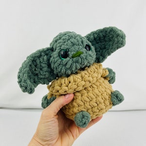 May include: A handmade crochet doll resembling a popular character. The doll has large, dark eyes, oversized ears, and a small green nose. It is mainly green and tan, with a soft, textured look. The doll is approximately 15 cm tall.