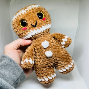 May include: A brown crocheted gingerbread man doll with white accents. The doll has large, glittery eyes, rosy cheeks, and a smiling face. White frosting-like trim outlines the head and limbs. Two white buttons adorn the body, and the doll is made of a soft, textured material.