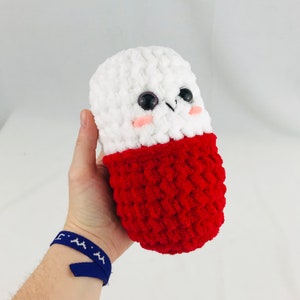 May include: A white and red crocheted pill-shaped plush toy with a cute face. The toy has black eyes, pink cheeks, and a small white nose.