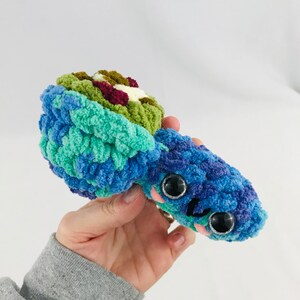May include: A crocheted stuffed toy in the shape of a sea creature with blue, green, and purple yarn. The toy has large black eyes and a small pink mouth.