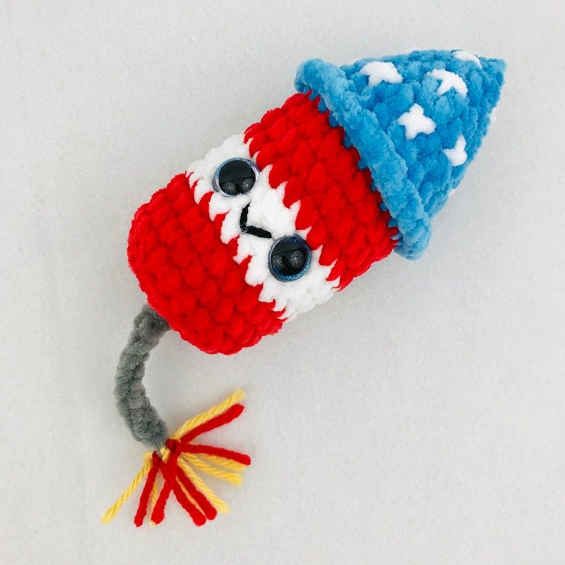 May include: A crocheted plush toy shaped like a firework with a red, white, and blue color scheme. The toy has googly eyes and a blue crocheted hat.