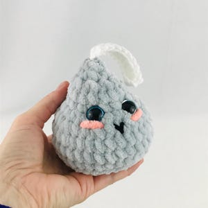 May include: A gray crocheted plush toy shaped like a teardrop with a white crocheted loop on top. The toy has large black eyes with blue pupils and pink cheeks.