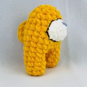 Puede incluir: Un juguete de peluche amarillo tejido a crochet con forma de personaje del videojuego Among Us. El juguete tiene una cara blanca con un contorno negro.