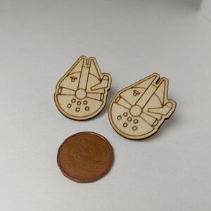 Puede incluir: Un par de pendientes de madera con forma de nave espacial Millennium Falcon de Star Wars. Los pendientes son de color marrón claro y tienen un diseño detallado.