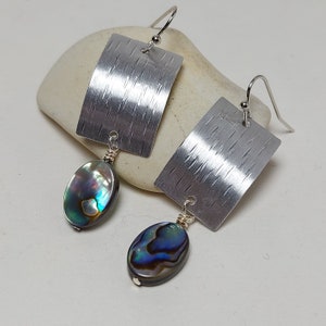 Può includere: Orecchini d'argento con un pezzo rettangolare in metallo testurizzato e un ciondolo a forma di lacrima in madreperla di abalone iridescente.