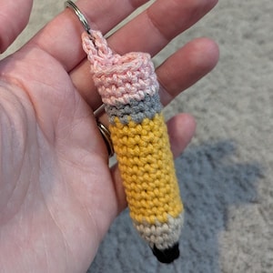 Pode incluir: Chaveiro de lápis de crochê com corpo amarelo, borracha cinza e ponta preta. A alça superior é rosa. O chaveiro é preso a um anel de metal prateado.