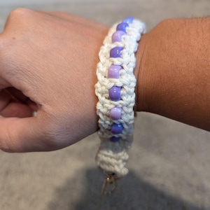May include: A white crocheted bracelet with a row of purple and lavender beads. The bracelet has a gold clasp.