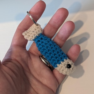 May include: A small, handmade crochet fish keychain featuring a blue body with cream-colored head and tail sections. The fish has a single black bead eye and is attached to a circular silver-toned metal split ring for keys or bag attachment.