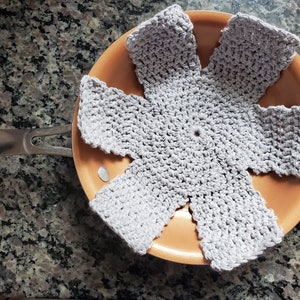 May include: A gray crocheted pot holder in the shape of a flower, placed inside a copper-colored pan.