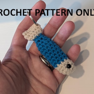 May include: A small crochet fish keychain featuring a blue body with beige head and tail sections, a single black bead eye, and a metal keyring attachment. The text CROCHET PATTERN ONLY is displayed in black at the top of the image.
