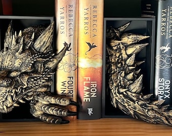 May include: A pair of dragon bookends made of a dark gray resin with gold accents. The dragon's head is on the left and the tail is on the right. The bookends are holding up two books with the titles "Iron Flame" and "Fourth Win" by Rebecca Yarros.
