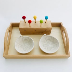 May include: A wooden tray with a light finish holds a wooden block with five colorful felt balls on wooden pegs. Two small, off-white rope bowls sit below. The tray has handles.