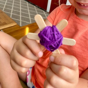 May include: A handmade craft project featuring a star-shaped design made from wooden popsicle sticks and wrapped with vibrant purple yarn. The craft is held in a person's hands, showcasing the DIY nature of the project.
