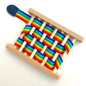 May include: A wooden weaving loom with a rainbow ribbon woven through it. The loom has a blue leather tab on the top.