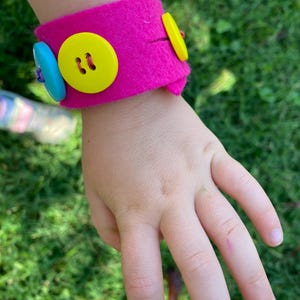 May include: A pink felt bracelet with yellow and blue buttons. The bracelet is on a child's wrist.
